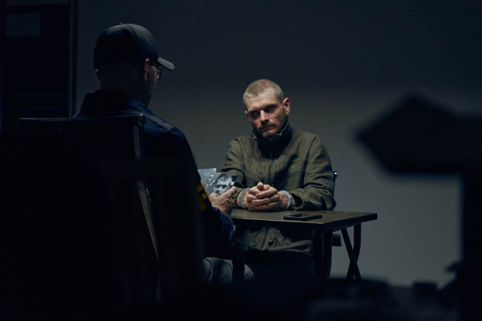 A serious man is being interviewed, presumably about a crime. He sits at a table with his hands clasped, facing a man in uniform.